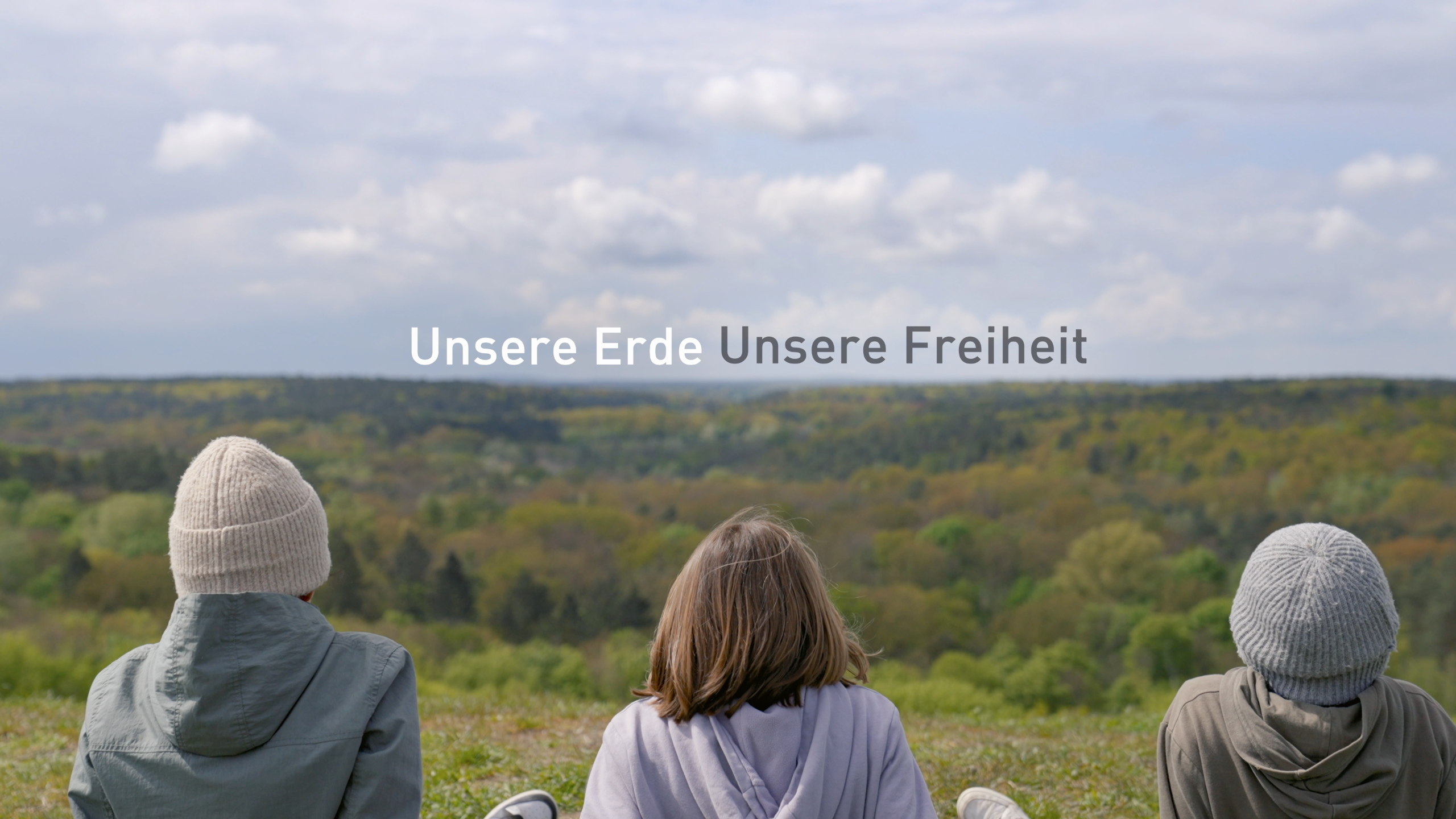 Drei Kinder sitzen auf einem Hügel und schauen über einen Wald. In der Mitte steht der Titel des Films, „Unsere Erde, Unsere Freiheit“.