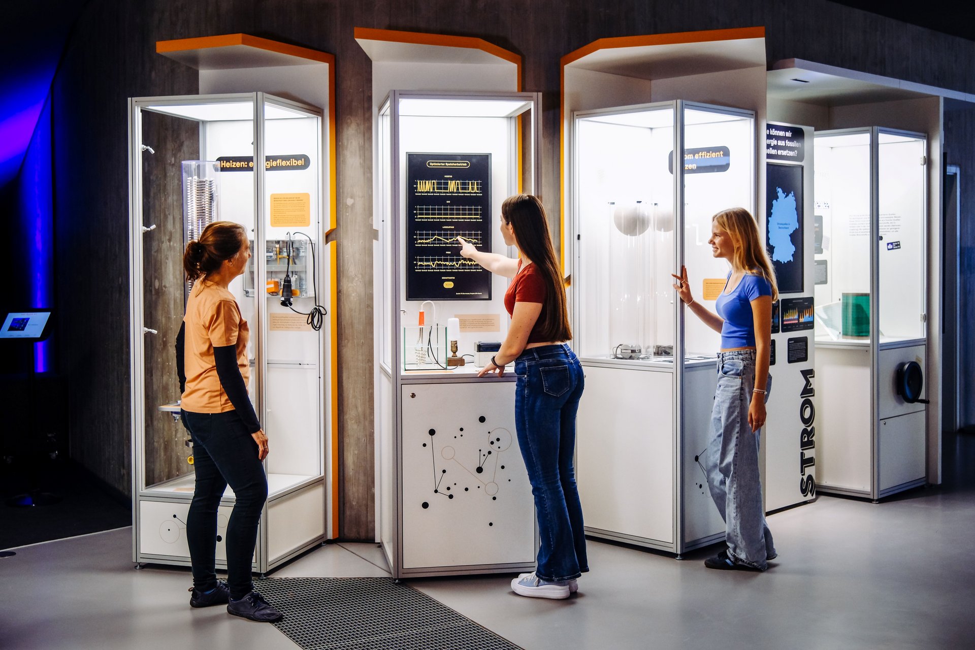 Drei junge Frauen stehen an Schaukästen einer Ausstellung. Eine von ihnen zeigt auf einen Grafen, der sich darin befindet.