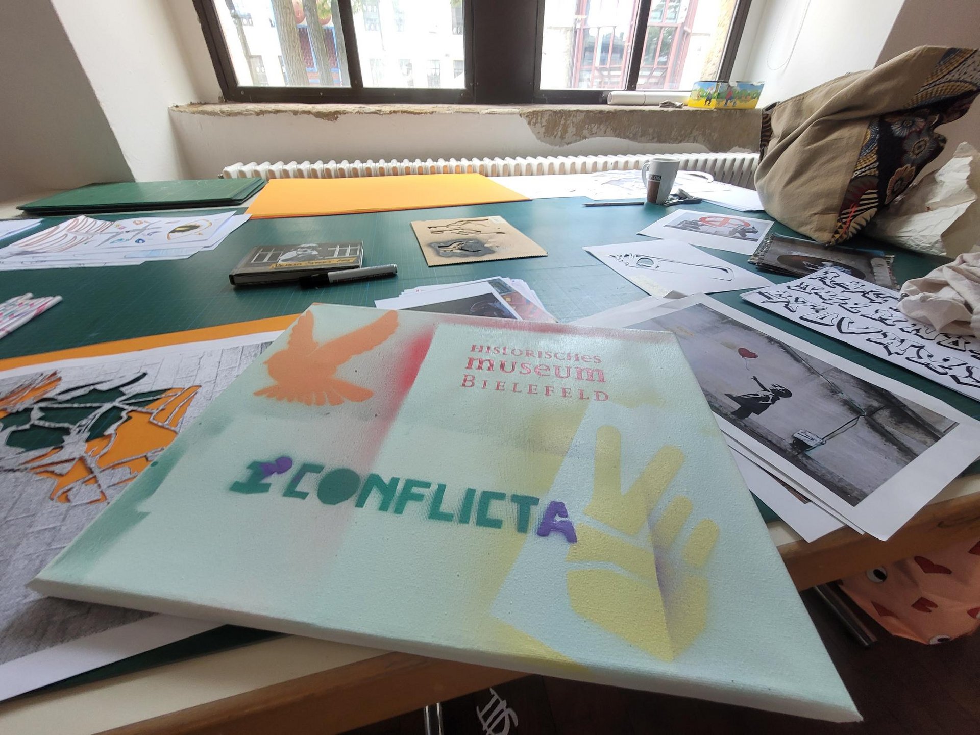Ein Tisch mit Bildern darauf. Auf eine Leinwand ist „Historisches Museum Bielefeld ConflictA“ gesprayed.
